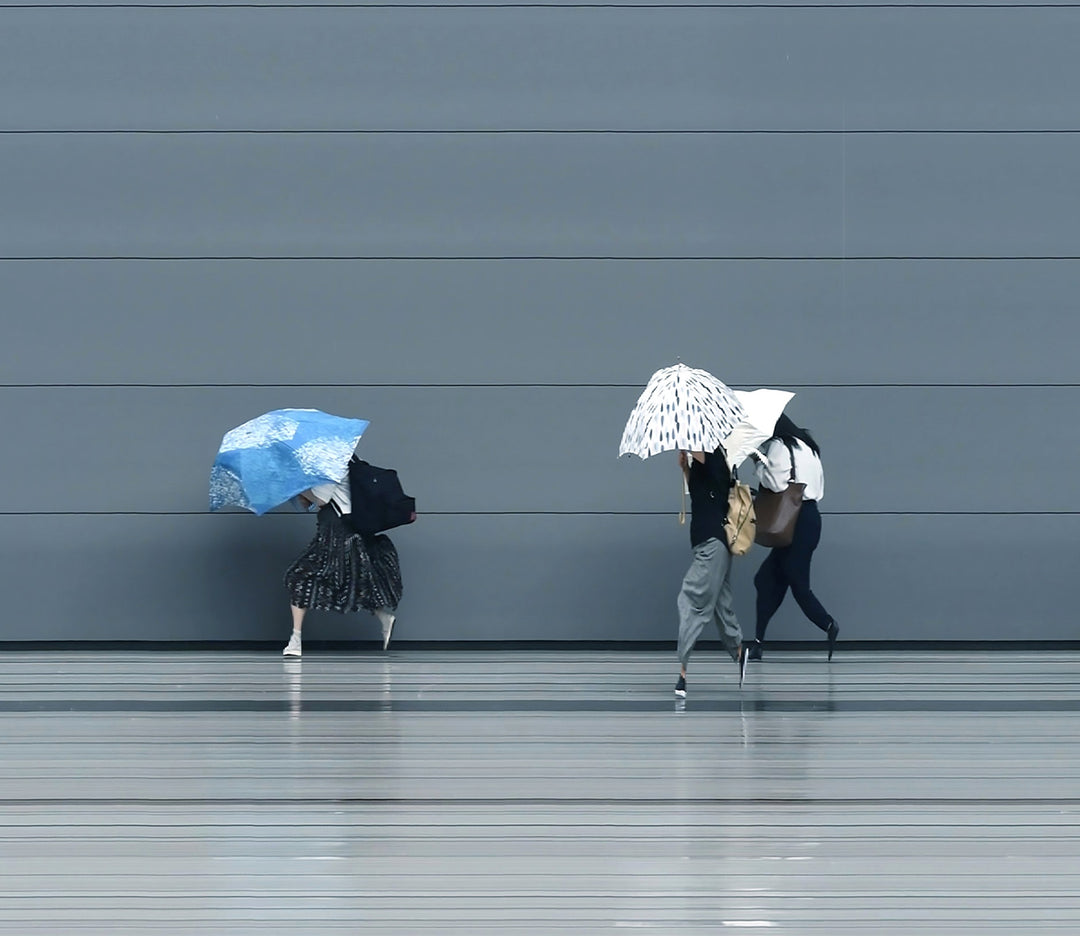 A stylised time-slice image of Tokyo commuters struggling against wind and rain during a summer storm. Each figure holds an umbrella as they move across a smooth, steel-grey background—some distorted mid-stride due to the artist’s high-speed video scan technique. The image captures both the uniformity and individuality of city life under pressure.