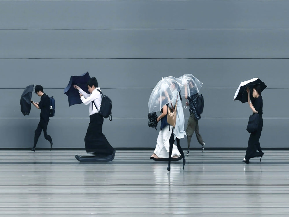A stylised time-slice image of Tokyo commuters struggling against wind and rain during a summer storm. Each figure holds an umbrella as they move across a smooth, steel-grey background—some distorted mid-stride due to the artist’s high-speed video scan technique. The image captures both the uniformity and individuality of city life under pressure.