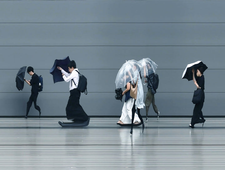 A stylised time-slice image of Tokyo commuters struggling against wind and rain during a summer storm. Each figure holds an umbrella as they move across a smooth, steel-grey background—some distorted mid-stride due to the artist’s high-speed video scan technique. The image captures both the uniformity and individuality of city life under pressure.