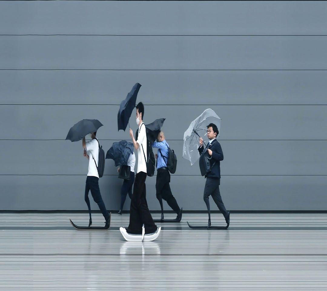 A stylised time-slice image of Tokyo commuters struggling against wind and rain during a summer storm. Each figure holds an umbrella as they move across a smooth, steel-grey background—some distorted mid-stride due to the artist’s high-speed video scan technique. The image captures both the uniformity and individuality of city life under pressure.