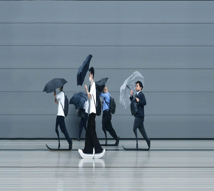 A stylised time-slice image of Tokyo commuters struggling against wind and rain during a summer storm. Each figure holds an umbrella as they move across a smooth, steel-grey background—some distorted mid-stride due to the artist’s high-speed video scan technique. The image captures both the uniformity and individuality of city life under pressure.