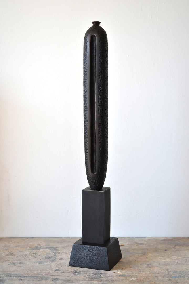 TORR by Nicola Beattie – tall, slender walnut sculpture with textured, rounded form and deep central vertical recess, mounted on a solid stepped base. Rich dark brown finish with natural grain, evoking ancient totems and minimalist modern design.