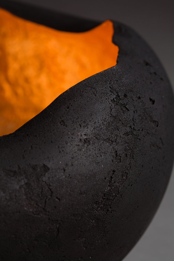 Tracy Nicholls glass sculpture Glow — a hand-formed pâte de verre orb with a rough matte black exterior and glowing copper leaf interior. The vessel radiates warmth, with light reflecting across its curved inner surface, creating the illusion of fire glowing from within.