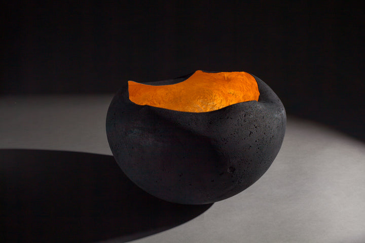 Tracy Nicholls glass sculpture Glow — a hand-formed pâte de verre orb with a rough matte black exterior and glowing copper leaf interior. The vessel radiates warmth, with light reflecting across its curved inner surface, creating the illusion of fire glowing from within.