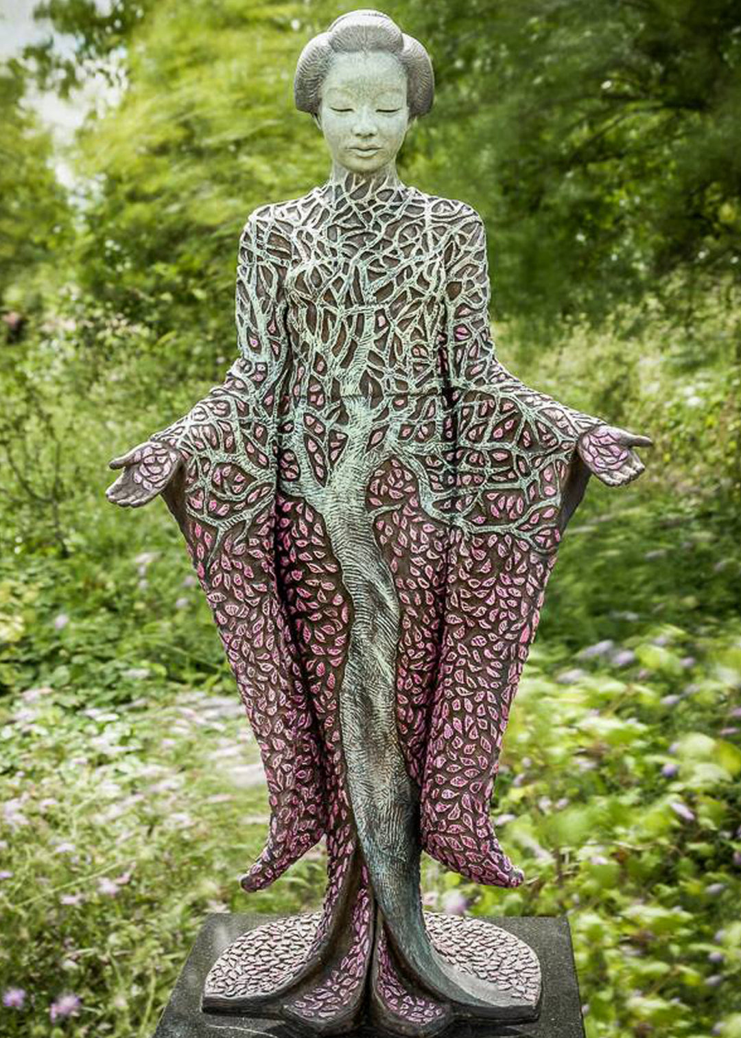 Bronze sculpture of a serene female figure standing with softly extended arms, her body intricately patterned as a blossoming tree. Pale green branches and trunk flow through her torso, while hundreds of delicate pink leaves form her robe and cascade toward the ground. The sculpture stands outdoors, surrounded by soft, blurred greenery that enhances its tranquil, meditative presence.