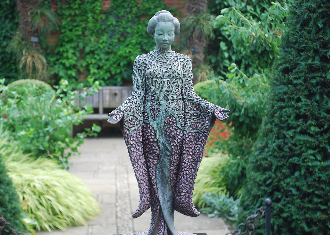 Bronze sculpture of a serene female figure standing with softly extended arms, her body intricately patterned as a blossoming tree. Pale green branches and trunk flow through her torso, while hundreds of delicate pink leaves form her robe and cascade toward the ground. The sculpture stands outdoors, surrounded by soft, blurred greenery that enhances its tranquil, meditative presence.