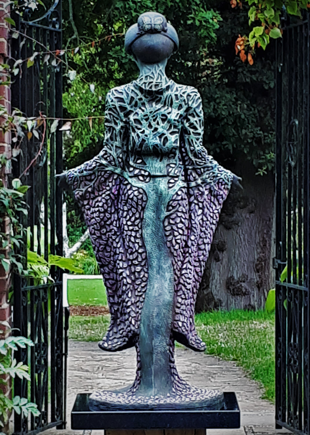 Bronze sculpture of a serene female figure standing with softly extended arms, her body intricately patterned as a blossoming tree. Pale green branches and trunk flow through her torso, while hundreds of delicate pink leaves form her robe and cascade toward the ground. The sculpture stands outdoors, surrounded by soft, blurred greenery that enhances its tranquil, meditative presence.