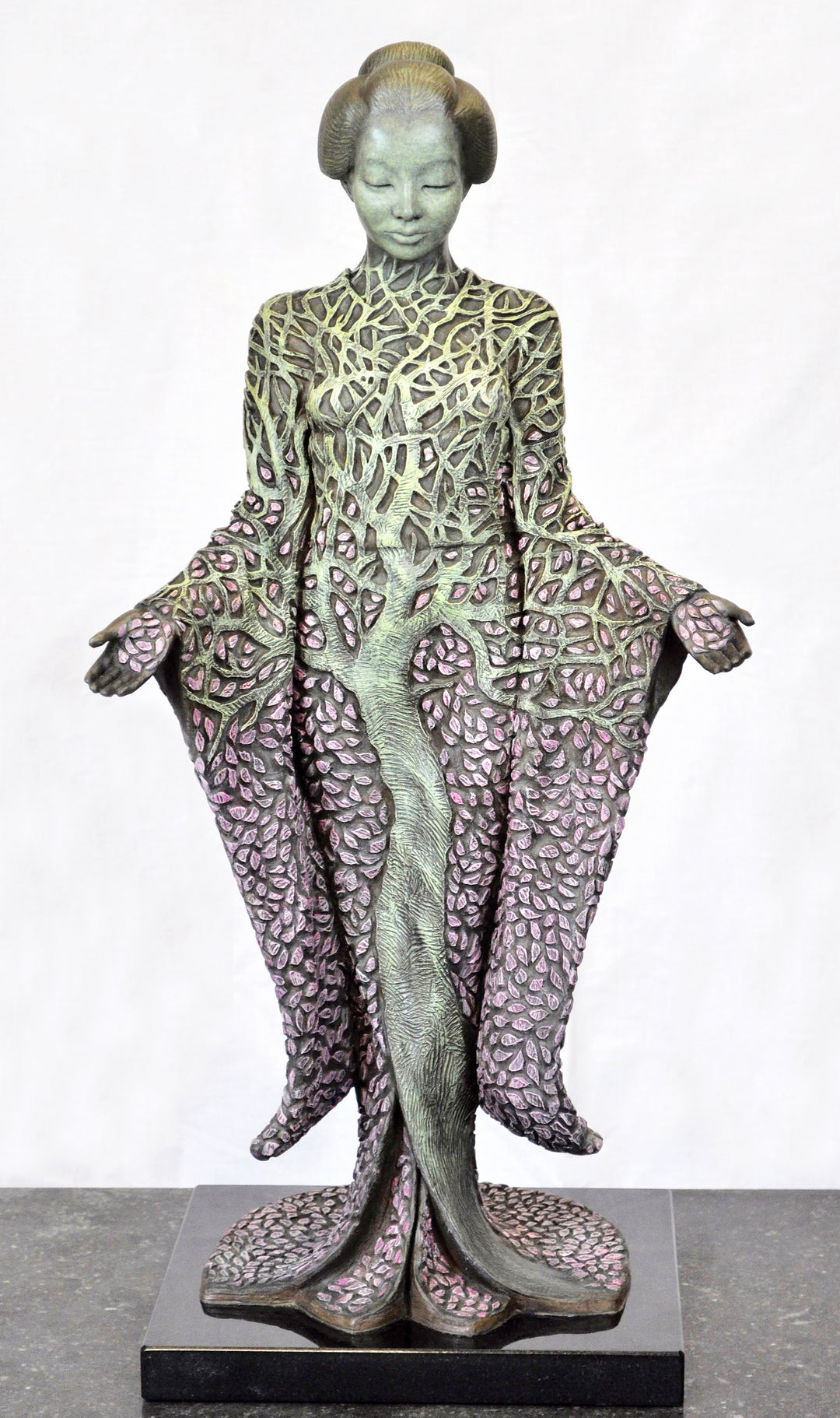 Bronze sculpture of a serene female figure standing with softly extended arms, her body intricately patterned as a blossoming tree. Pale green branches and trunk flow through her torso, while hundreds of delicate pink leaves form her robe and cascade toward the ground. The sculpture stands outdoors, surrounded by soft, blurred greenery that enhances its tranquil, meditative presence.