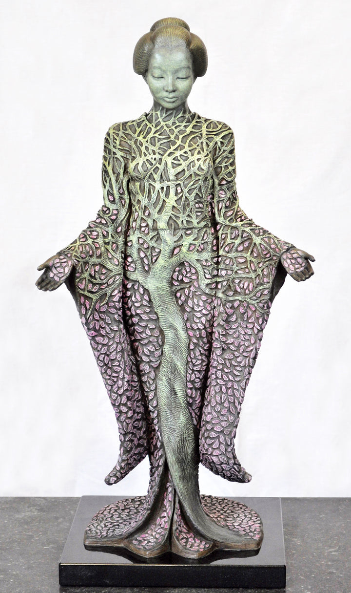 Bronze sculpture of a serene female figure standing with softly extended arms, her body intricately patterned as a blossoming tree. Pale green branches and trunk flow through her torso, while hundreds of delicate pink leaves form her robe and cascade toward the ground. The sculpture stands outdoors, surrounded by soft, blurred greenery that enhances its tranquil, meditative presence.