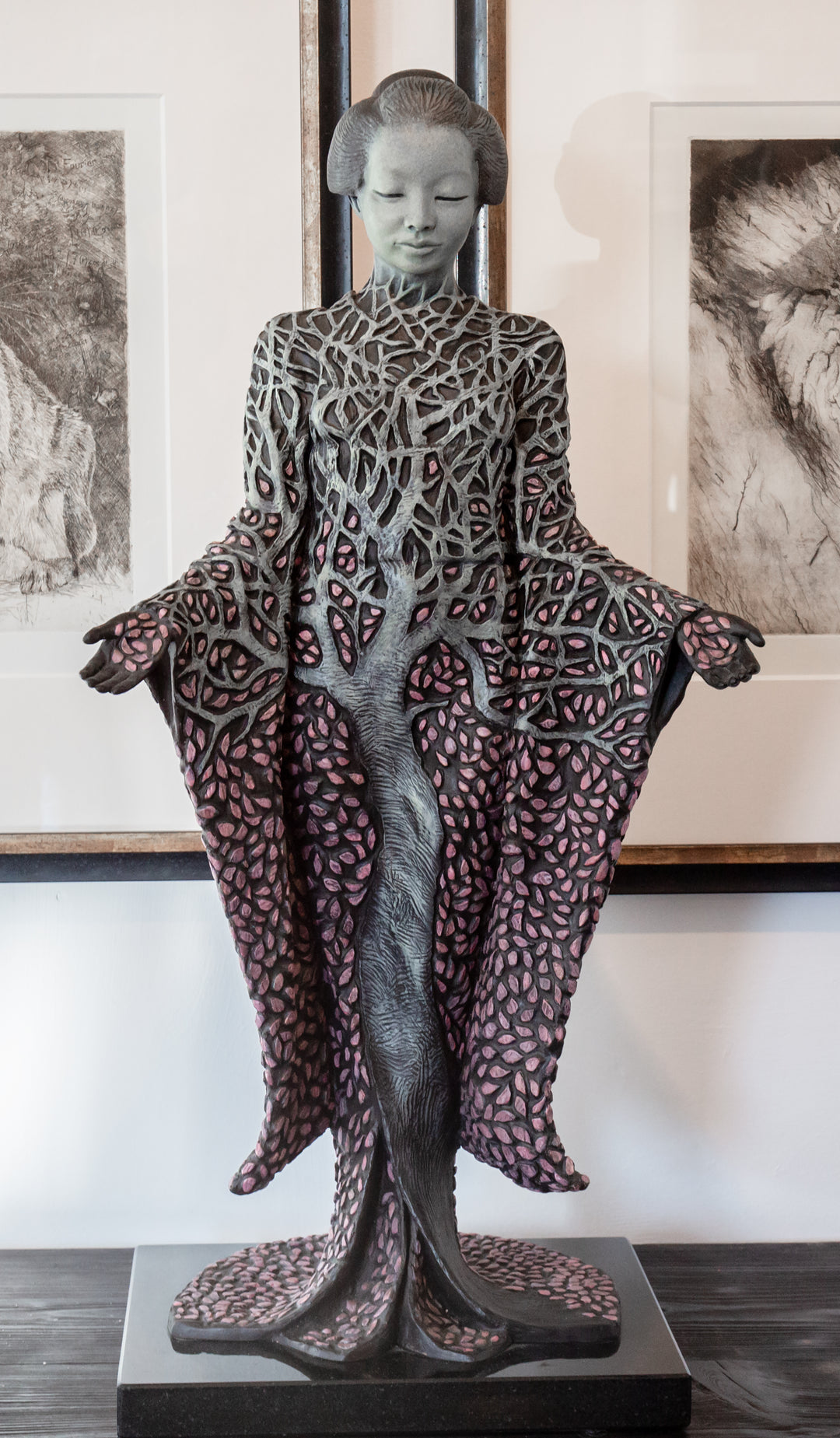 Bronze sculpture of a serene female figure standing with softly extended arms, her body intricately patterned as a blossoming tree. Pale green branches and trunk flow through her torso, while hundreds of delicate pink leaves form her robe and cascade toward the ground. The sculpture stands outdoors, surrounded by soft, blurred greenery that enhances its tranquil, meditative presence.