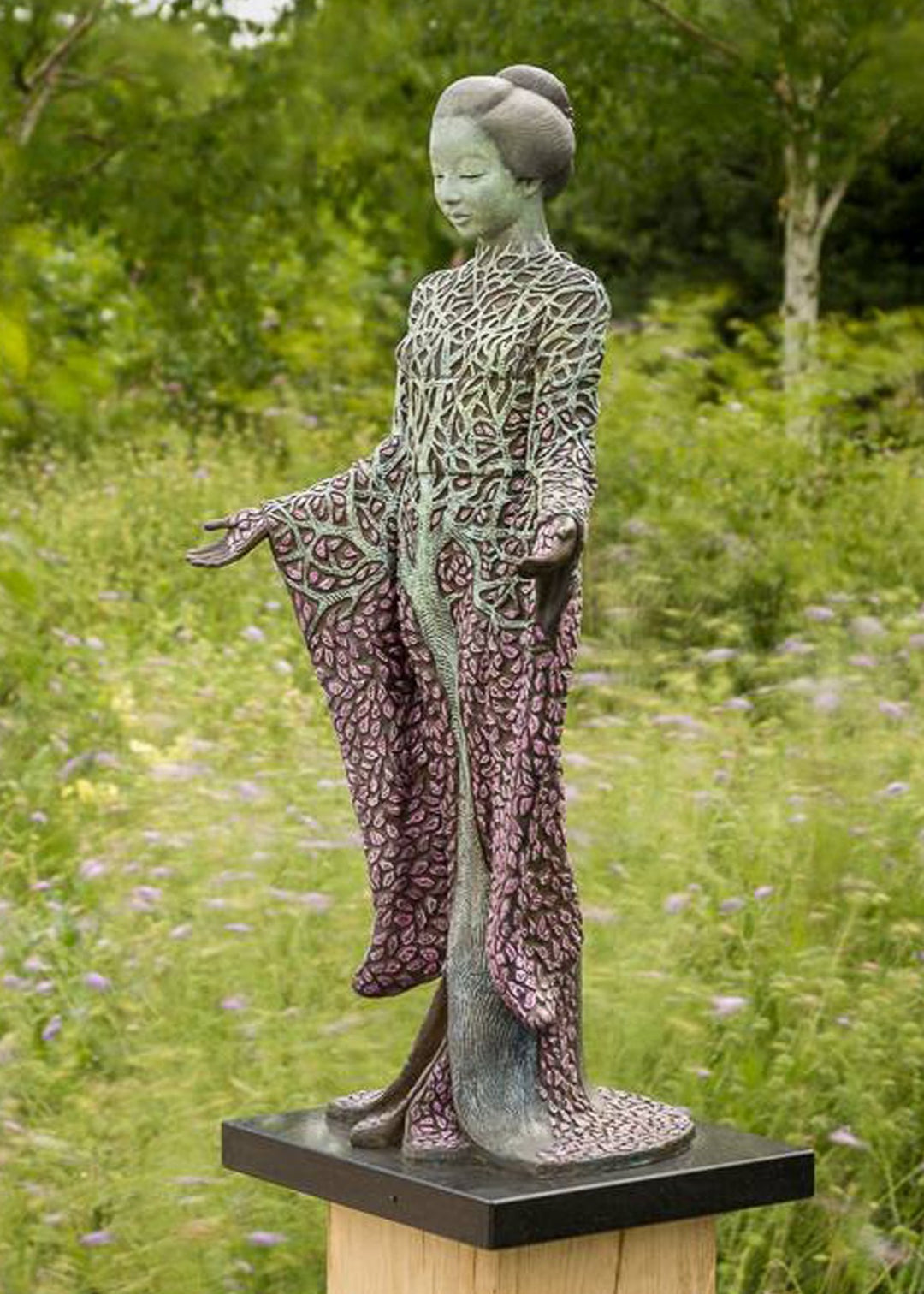 Bronze sculpture of a serene female figure standing with softly extended arms, her body intricately patterned as a blossoming tree. Pale green branches and trunk flow through her torso, while hundreds of delicate pink leaves form her robe and cascade toward the ground. The sculpture stands outdoors, surrounded by soft, blurred greenery that enhances its tranquil, meditative presence.
