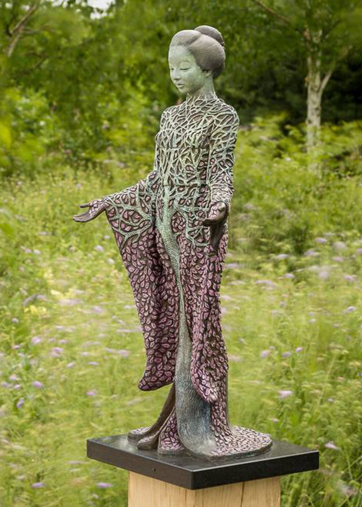 Bronze sculpture of a serene female figure standing with softly extended arms, her body intricately patterned as a blossoming tree. Pale green branches and trunk flow through her torso, while hundreds of delicate pink leaves form her robe and cascade toward the ground. The sculpture stands outdoors, surrounded by soft, blurred greenery that enhances its tranquil, meditative presence.