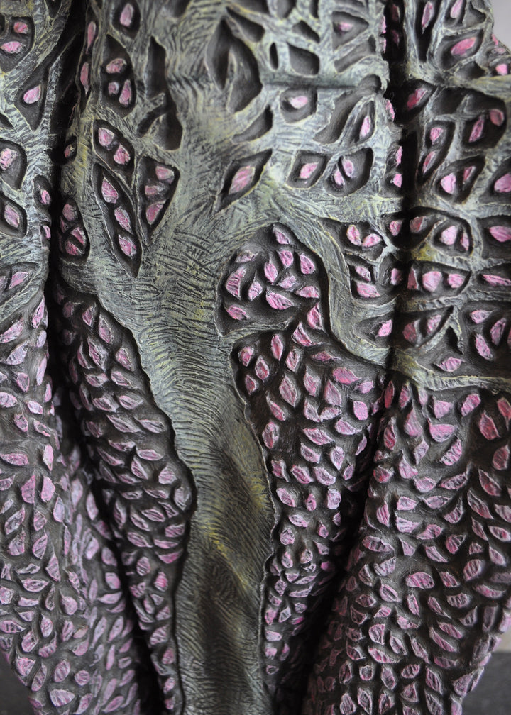 Bronze sculpture of a serene female figure standing with softly extended arms, her body intricately patterned as a blossoming tree. Pale green branches and trunk flow through her torso, while hundreds of delicate pink leaves form her robe and cascade toward the ground. The sculpture stands outdoors, surrounded by soft, blurred greenery that enhances its tranquil, meditative presence.
