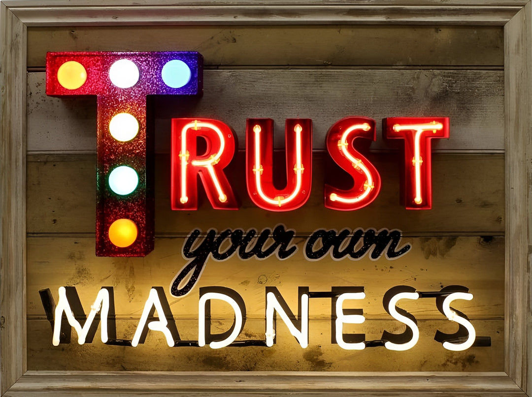 Framed neon artwork by Matthew Bracey reading “Trust your own MADNESS.” The word “TRUST” is bold in red neon, with a glittered letter ‘T’ lit by colourful carnival-style bulbs. “Your own” is rendered in a black cursive style, and “MADNESS” shines in bright white neon, all set against a distressed wooden panel backdrop.