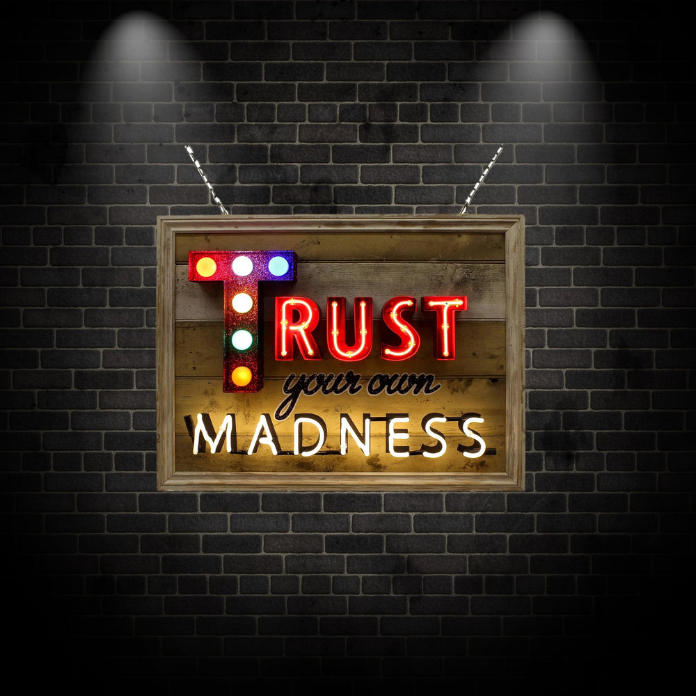 Framed neon artwork by Matthew Bracey reading “Trust your own MADNESS.” The word “TRUST” is bold in red neon, with a glittered letter ‘T’ lit by colourful carnival-style bulbs. “Your own” is rendered in a black cursive style, and “MADNESS” shines in bright white neon, all set against a distressed wooden panel backdrop.