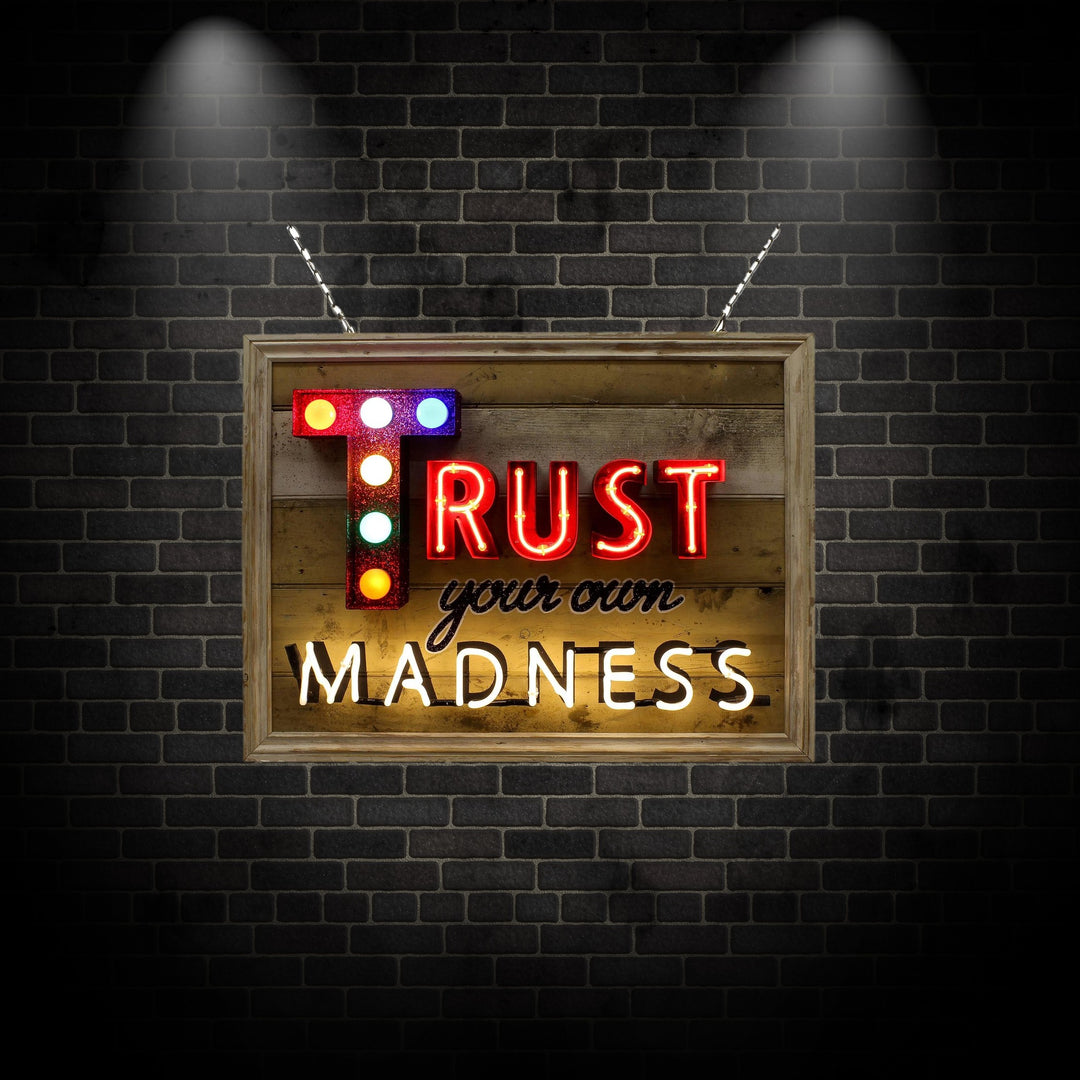 Framed neon artwork by Matthew Bracey reading “Trust your own MADNESS.” The word “TRUST” is bold in red neon, with a glittered letter ‘T’ lit by colourful carnival-style bulbs. “Your own” is rendered in a black cursive style, and “MADNESS” shines in bright white neon, all set against a distressed wooden panel backdrop.