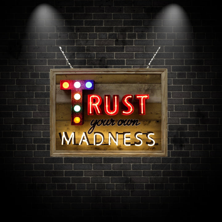 Framed neon artwork by Matthew Bracey reading “Trust your own MADNESS.” The word “TRUST” is bold in red neon, with a glittered letter ‘T’ lit by colourful carnival-style bulbs. “Your own” is rendered in a black cursive style, and “MADNESS” shines in bright white neon, all set against a distressed wooden panel backdrop.