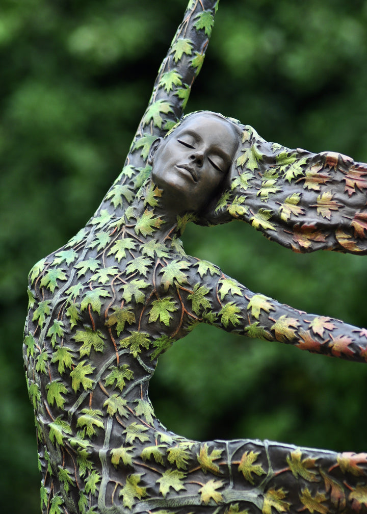 Bronze sculpture of a female figure balancing gracefully on one foot, her body and flowing dress covered in hand-painted maple leaves that shift from fresh greens to warm yellows, oranges and deep autumnal reds. She leans into the wind with one arm raised, hair swept back, standing outdoors against dense green foliage.