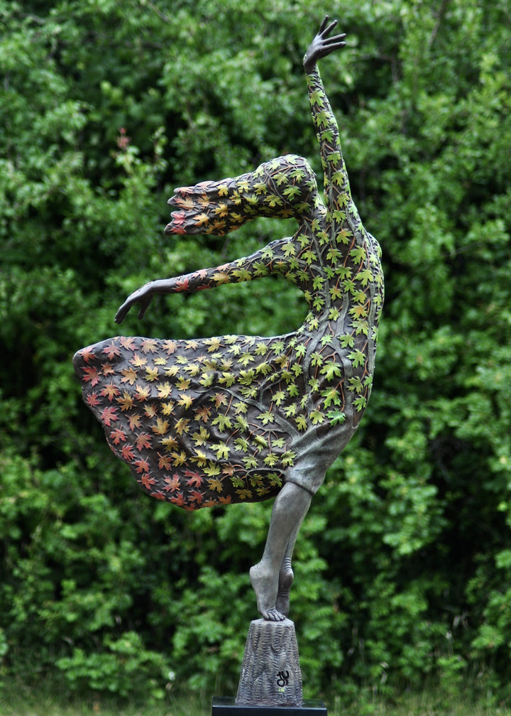 Bronze sculpture of a female figure balancing gracefully on one foot, her body and flowing dress covered in hand-painted maple leaves that shift from fresh greens to warm yellows, oranges and deep autumnal reds. She leans into the wind with one arm raised, hair swept back, standing outdoors against dense green foliage.