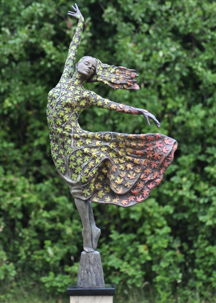Bronze sculpture of a female figure balancing gracefully on one foot, her body and flowing dress covered in hand-painted maple leaves that shift from fresh greens to warm yellows, oranges and deep autumnal reds. She leans into the wind with one arm raised, hair swept back, standing outdoors against dense green foliage.