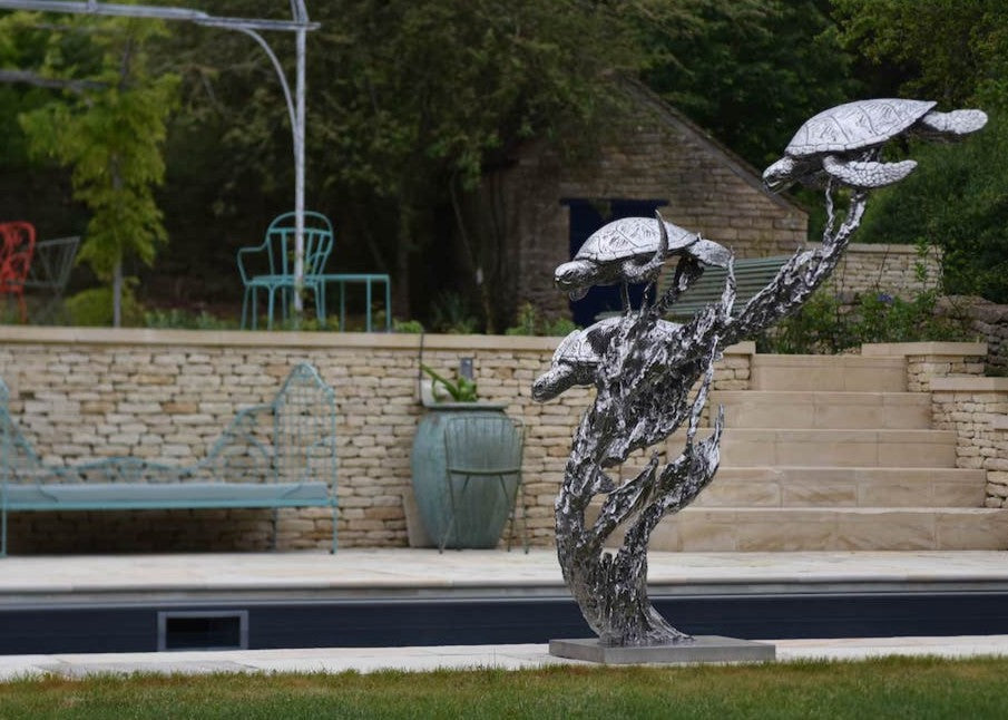 ‘Turtles Stainless Steel’ by Hamish Mackie, a limited-edition marine-grade stainless steel wildlife sculpture depicting three life-size sea turtles swimming through an intricate arrangement of underwater foliage. The polished silver-grey finish highlights the turtles' detailed shells, textured flippers and graceful movement, while the sweeping, branching forms create a sense of flowing water. Mounted on a rectangular stainless steel base, this contemporary sculpture is suitable for indoor or outdoor display