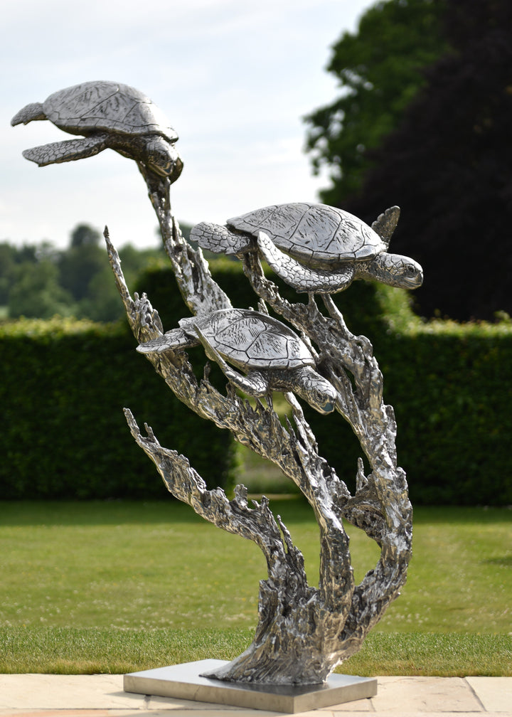 ‘Turtles Stainless Steel’ by Hamish Mackie, a limited-edition marine-grade stainless steel wildlife sculpture depicting three life-size sea turtles swimming through an intricate arrangement of underwater foliage. The polished silver-grey finish highlights the turtles' detailed shells, textured flippers and graceful movement, while the sweeping, branching forms create a sense of flowing water. Mounted on a rectangular stainless steel base, this contemporary sculpture is suitable for indoor or outdoor display