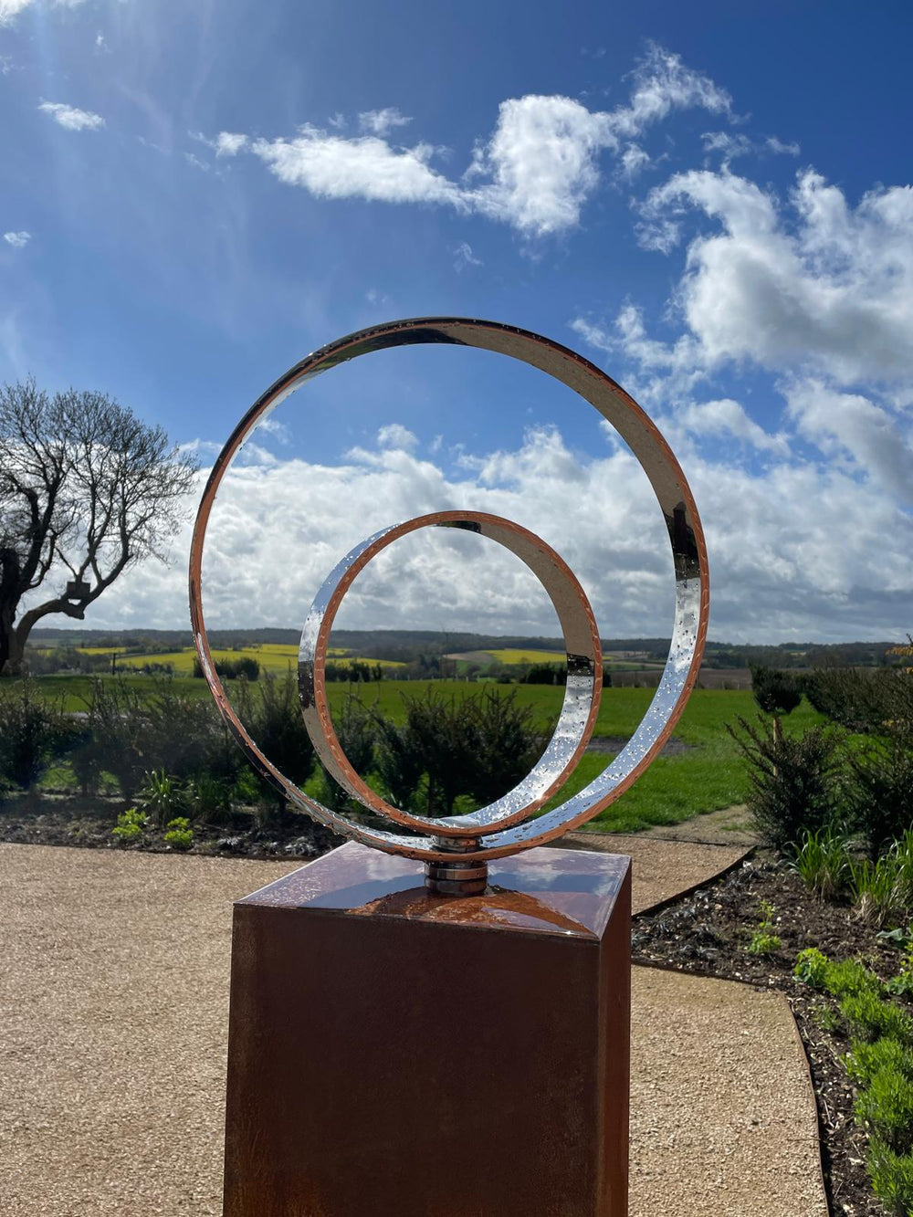 Polished stainless steel kinetic concentric ring sculpture by Tom Hiscox, mounted on a tall corten steel plinth and set against a sweeping rural landscape with open skies and distant fields.