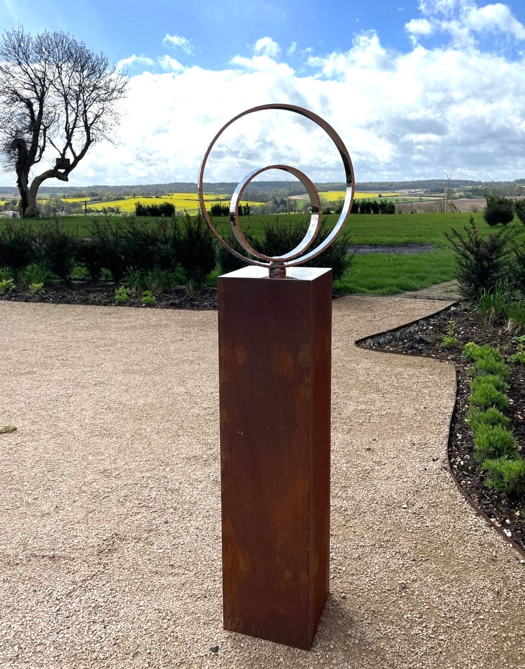 Polished stainless steel kinetic concentric ring sculpture by Tom Hiscox, mounted on a tall corten steel plinth and set against a sweeping rural landscape with open skies and distant fields.