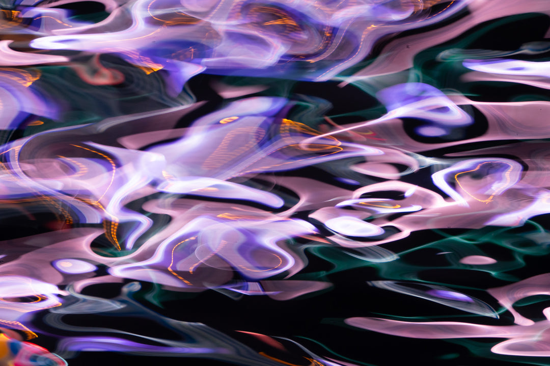 Abstract photograph capturing soft violet, pink, and electric blue light reflections across the water's surface, creating a dreamy, fluid composition with hints of green and orange.