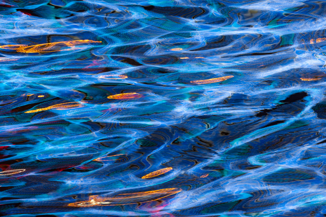 Abstract photograph of rippling water illuminated in vivid electric blues with streaks of amber, gold, turquoise, and occasional crimson accents, creating a mesmerising, painterly surface effect.