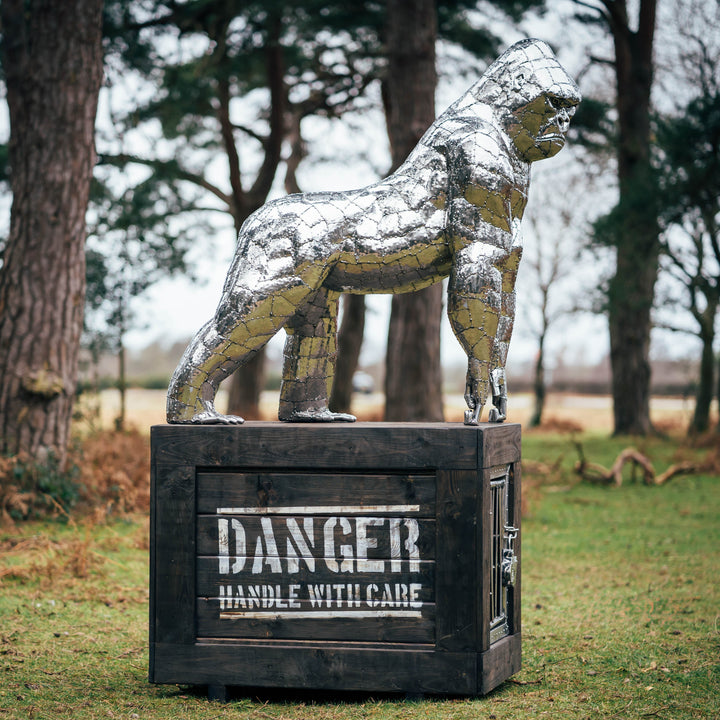 ‘Gorilla’ by Michael Turner, a unique, one-of-one stainless steel wildlife sculpture depicting a powerful silverback gorilla standing on all fours atop a large, dark wooden transport cage. Handcrafted from individually shaped and welded stainless steel panels, the sculpture features a polished silver finish, muscular shoulders, powerful limbs and intricately detailed facial features. The rustic timber cage displays bold white ‘DANGER - HANDLE WITH CARE’ lettering and a metal-barred hatch, adding humour and 