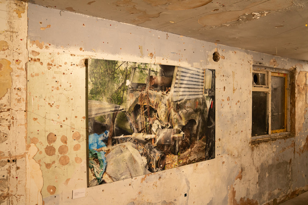A fragmented, semi-abstract composition of a crumpled van enveloped in rust, chains, and corroded metal, set against a backdrop of overgrown woodland. The surface of the vehicle is a patchwork of peeling paint and oxidised textures, with a twisted exhaust pipe and exposed mechanical parts jutting forward. Faint reflections and layered overlays create a painterly, dreamlike distortion across the entire scene.