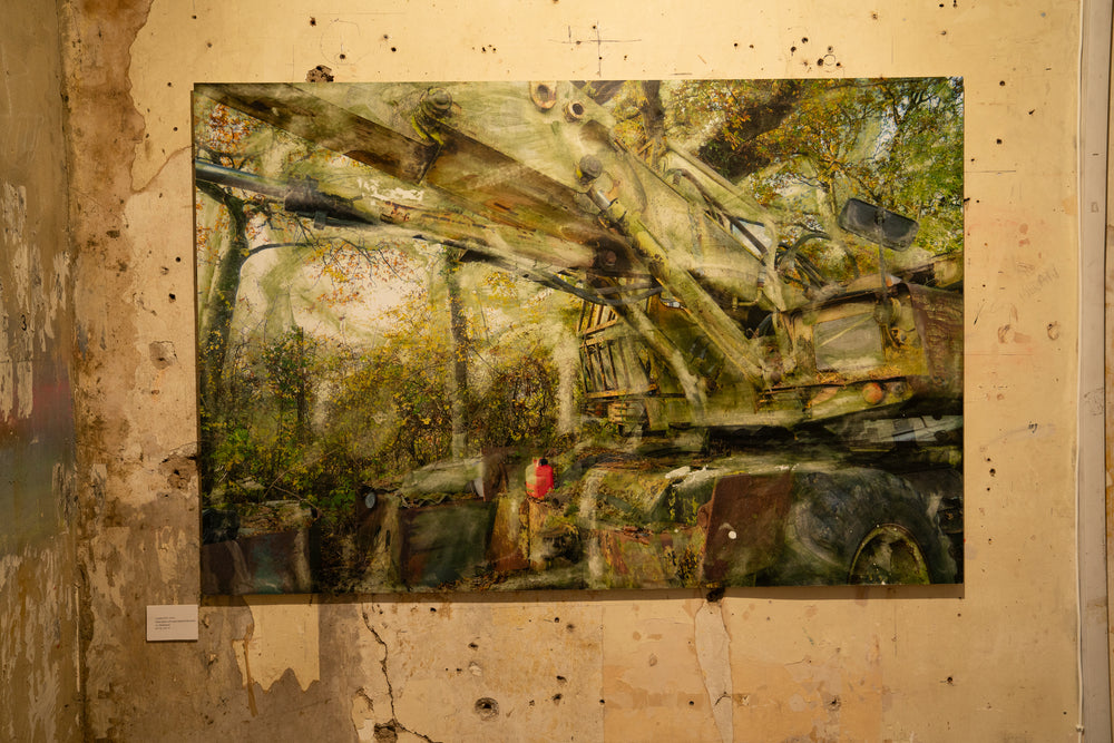 A decaying military-green crane or heavy machinery vehicle sits partially buried in a woodland clearing, its massive hydraulic arms rusted and streaked with oxidised residue. Autumnal foliage surrounds and overlays the scene, while painterly textures and ghosted transparencies give the piece a spectral, timeworn atmosphere. A solitary red fuel container adds a flash of colour against the muted industrial palette.