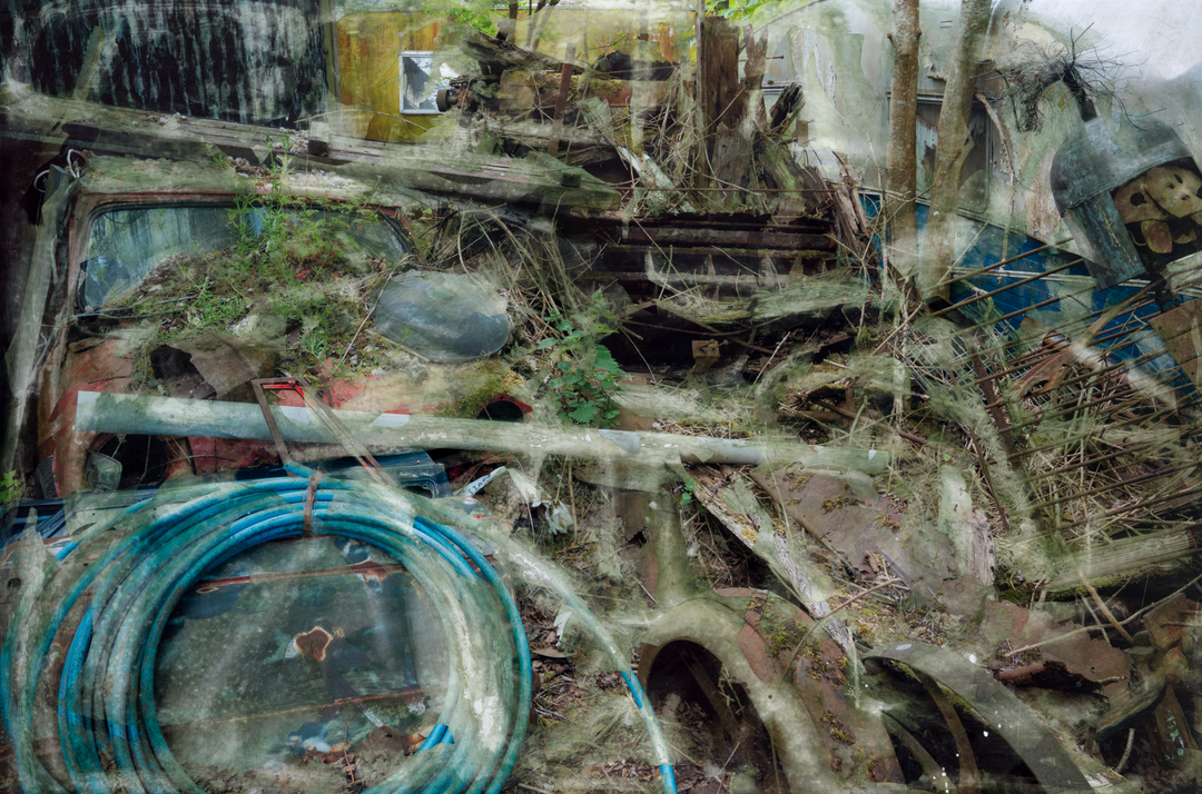 A densely layered, textural composition showing an entangled heap of decaying wood, metal, and automotive debris partially reclaimed by moss and undergrowth. Coiled blue piping, a fragmented red vehicle, and collapsing wooden structures are enveloped by soft overlays and distressed surfaces. The entire scene feels submerged in a dreamlike mist of erosion, time, and industrial memory.