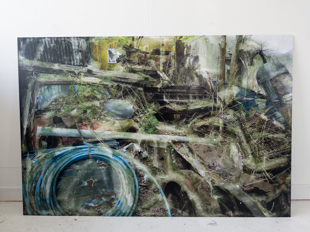 A densely layered, textural composition showing an entangled heap of decaying wood, metal, and automotive debris partially reclaimed by moss and undergrowth. Coiled blue piping, a fragmented red vehicle, and collapsing wooden structures are enveloped by soft overlays and distressed surfaces. The entire scene feels submerged in a dreamlike mist of erosion, time, and industrial memory.