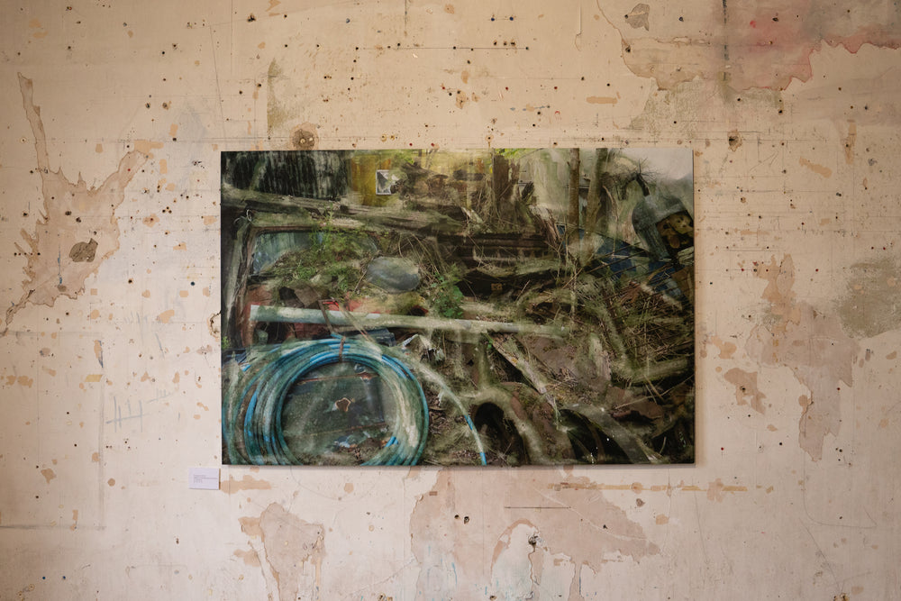 A densely layered, textural composition showing an entangled heap of decaying wood, metal, and automotive debris partially reclaimed by moss and undergrowth. Coiled blue piping, a fragmented red vehicle, and collapsing wooden structures are enveloped by soft overlays and distressed surfaces. The entire scene feels submerged in a dreamlike mist of erosion, time, and industrial memory.