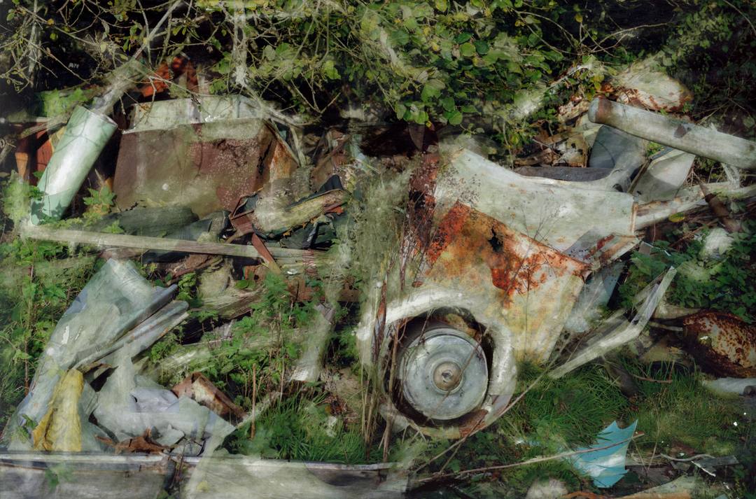A rich, layered image of industrial remnants and overgrown vegetation, where rust-streaked white vehicle panels, twisted metal, and collapsed timber are partially engulfed by brambles and wild greenery. A circular wheel hub juts from the foreground, surrounded by painterly overlays and textural transparency. The composition speaks to entropy, reclaiming nature, and the slow poetry of decay.