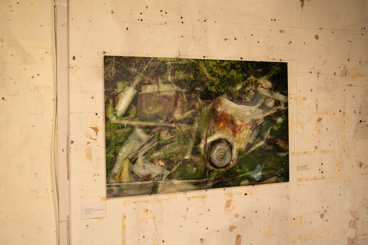 A rich, layered image of industrial remnants and overgrown vegetation, where rust-streaked white vehicle panels, twisted metal, and collapsed timber are partially engulfed by brambles and wild greenery. A circular wheel hub juts from the foreground, surrounded by painterly overlays and textural transparency. The composition speaks to entropy, reclaiming nature, and the slow poetry of decay.