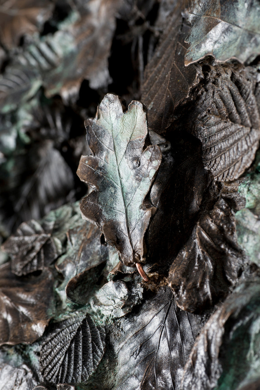 Tall patinated bronze sculpture of a female figure in a windswept, forward-leaning pose, emerging from a flowing base textured with detailed leaves. Her closed eyes and serene expression are partially obscured by sculpted foliage, symbolising a deep connection to nature and continual transformation. The figure’s dynamic form and elegant upward movement evoke renewal, seasonal change, and personal evolution. Rich bronze tones are enhanced by subtle green patination across the leafwork.