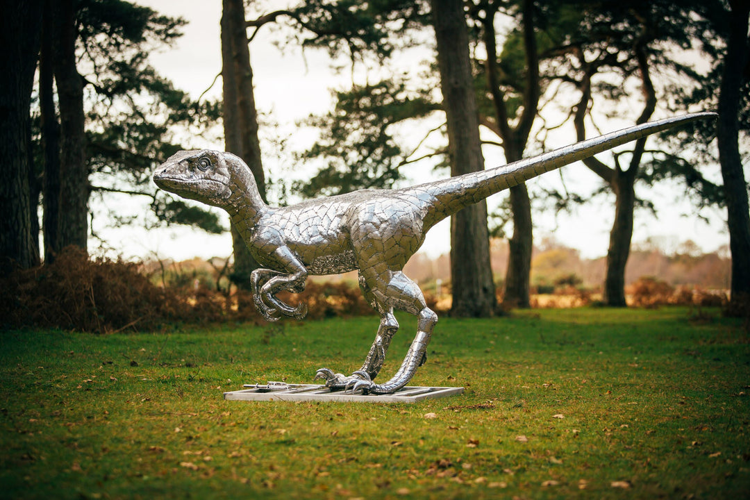 ‘Velociraptor’ by Michael Turner, a unique, one-of-one stainless steel dinosaur sculpture depicting a powerful velociraptor in an alert, dynamic pose. Handcrafted from intricately shaped and welded stainless steel, the sculpture features a polished silver finish, individually detailed metallic scales, muscular hind legs, sharp curved claws, smaller forelimbs and a long, sweeping tail. Standing on a rectangular stainless steel base, this large-scale contemporary sculpture captures the agility and predatory c