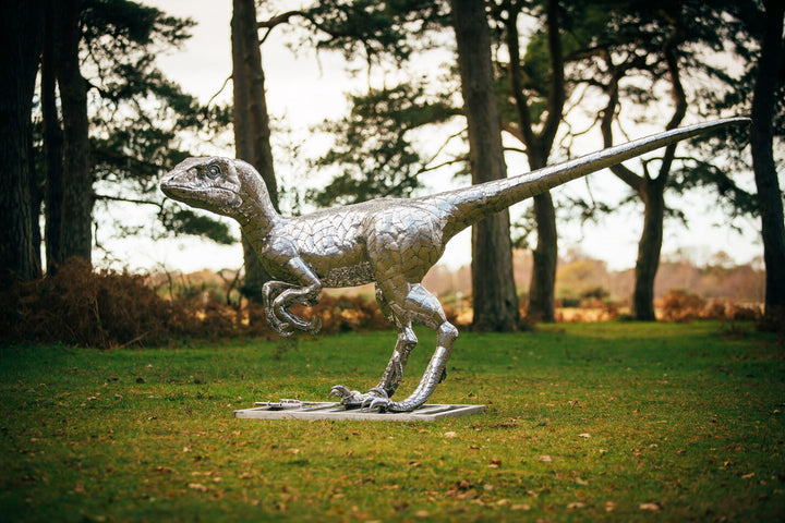 ‘Velociraptor’ by Michael Turner, a unique, one-of-one stainless steel dinosaur sculpture depicting a powerful velociraptor in an alert, dynamic pose. Handcrafted from intricately shaped and welded stainless steel, the sculpture features a polished silver finish, individually detailed metallic scales, muscular hind legs, sharp curved claws, smaller forelimbs and a long, sweeping tail. Standing on a rectangular stainless steel base, this large-scale contemporary sculpture captures the agility and predatory c