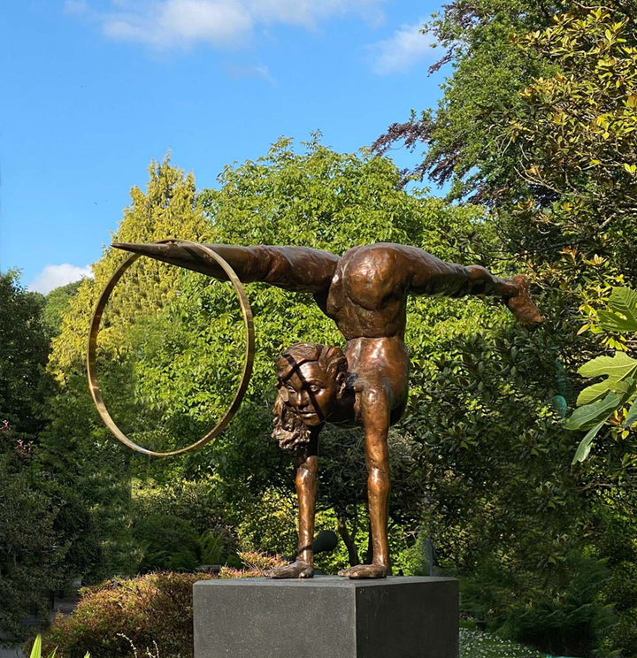 Teresa Wells’ Viktoriya (2018), limited edition bronze and brass sculpture on granite, portraying a powerful female acrobat in a one-armed handstand with legs extended horizontally, one foot tapering into a polished brass point that pierces a large brass hoop. The patinated bronze figure features finely modelled musculature, braided hair, and a dynamic forward gaze, balancing strength and grace. Mounted on a solid black granite base, the 100cm×30cm×50cm, 73kg piece is designed for impactful indoor display.