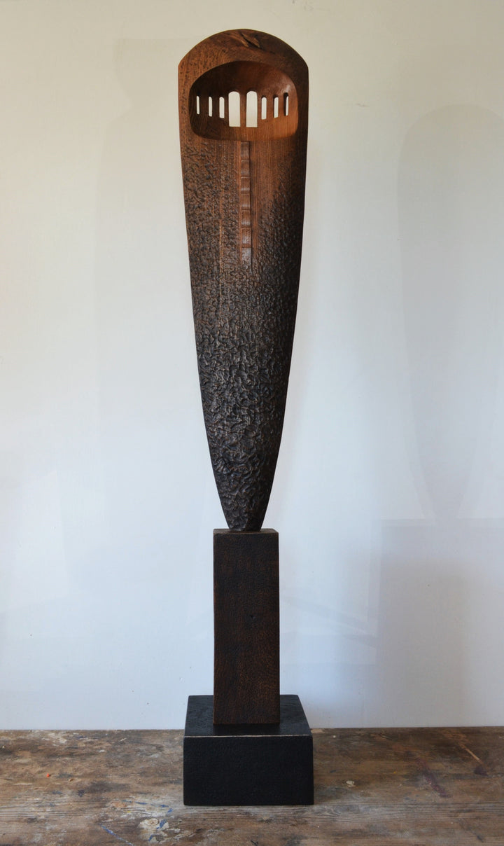 “Visori” by Nicola Beattie – a tall elm sculpture on an oak base, hand-carved with textured surface and visor-like openings. Part of her Blade series, it symbolises endurance, conflict, and the emergence of light from darkness.
