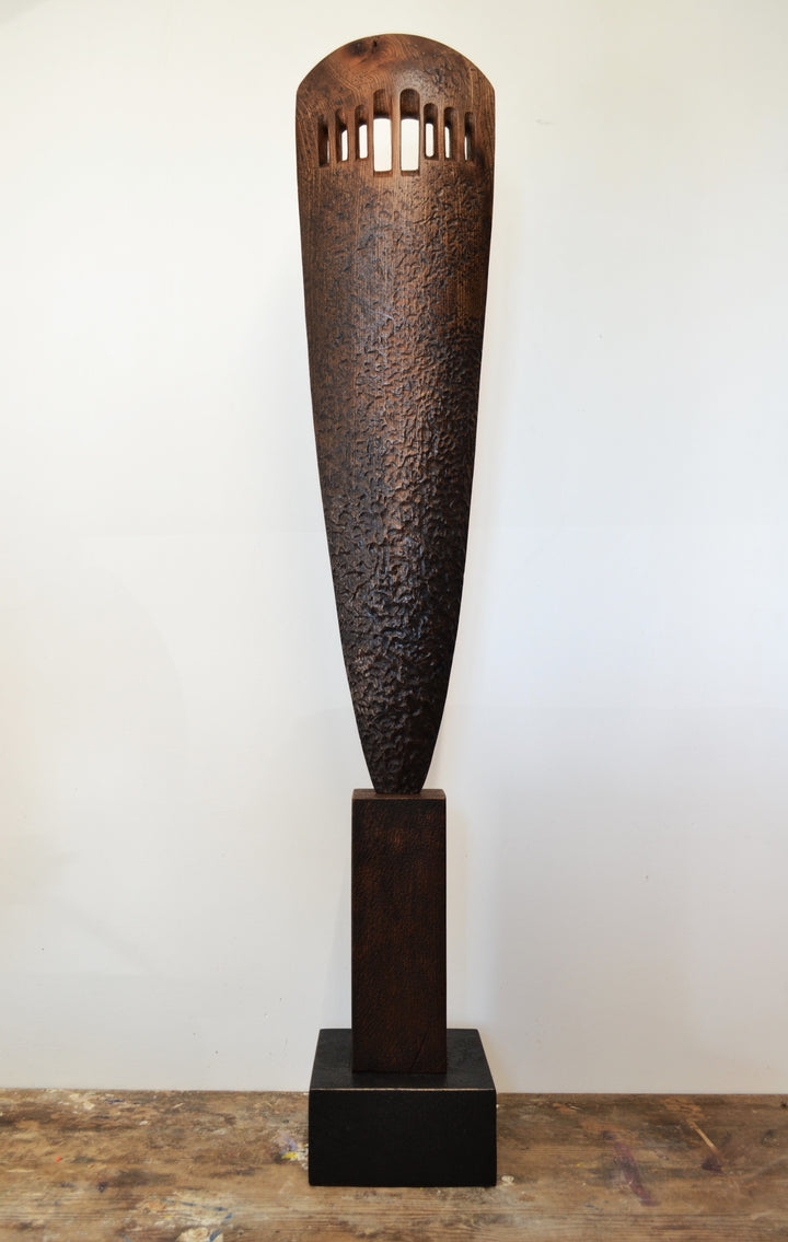 “Visori” by Nicola Beattie – a tall elm sculpture on an oak base, hand-carved with textured surface and visor-like openings. Part of her Blade series, it symbolises endurance, conflict, and the emergence of light from darkness.