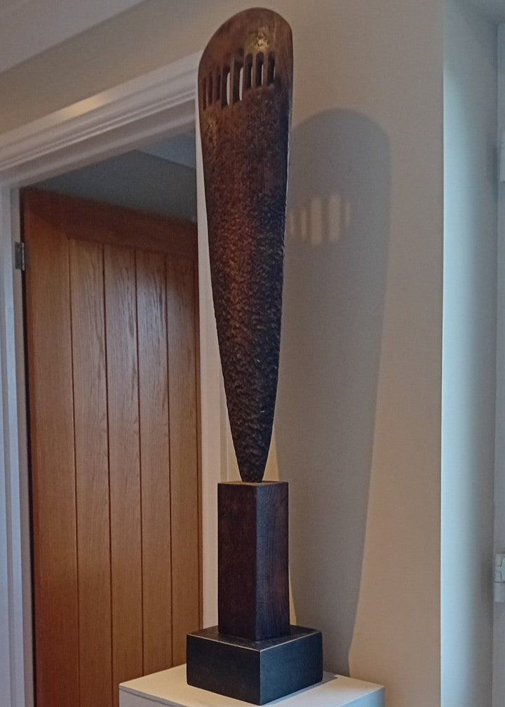 “Visori” by Nicola Beattie – a tall elm sculpture on an oak base, hand-carved with textured surface and visor-like openings. Part of her Blade series, it symbolises endurance, conflict, and the emergence of light from darkness.