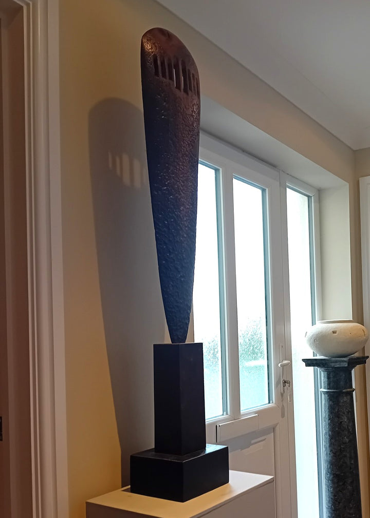 “Visori” by Nicola Beattie – a tall elm sculpture on an oak base, hand-carved with textured surface and visor-like openings. Part of her Blade series, it symbolises endurance, conflict, and the emergence of light from darkness.