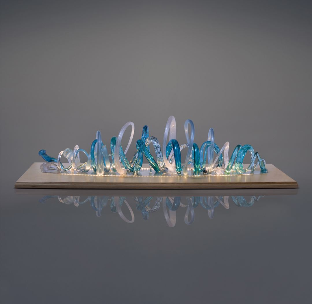 Glass sculpture of a wave, featuring flowing, ribbon-like forms in blue, aqua, and clear tones, set on a light wooden base.
