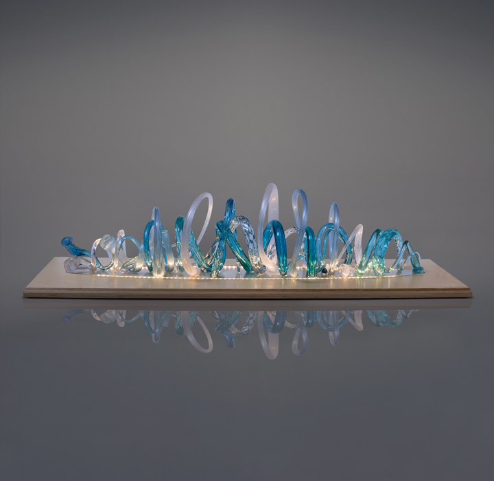 Glass sculpture of a wave, featuring flowing, ribbon-like forms in blue, aqua, and clear tones, set on a light wooden base.