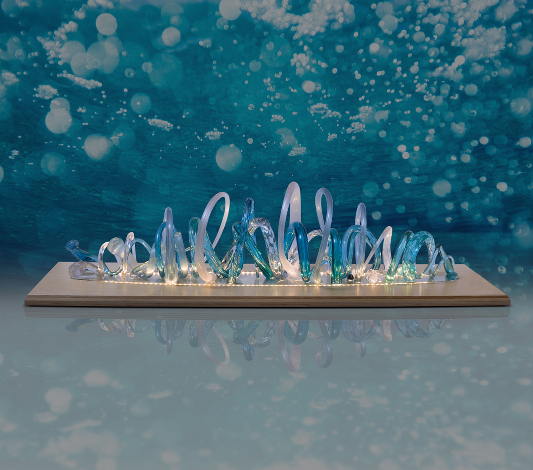 Glass sculpture of a wave, featuring flowing, ribbon-like forms in blue, aqua, and clear tones, set on a light wooden base.