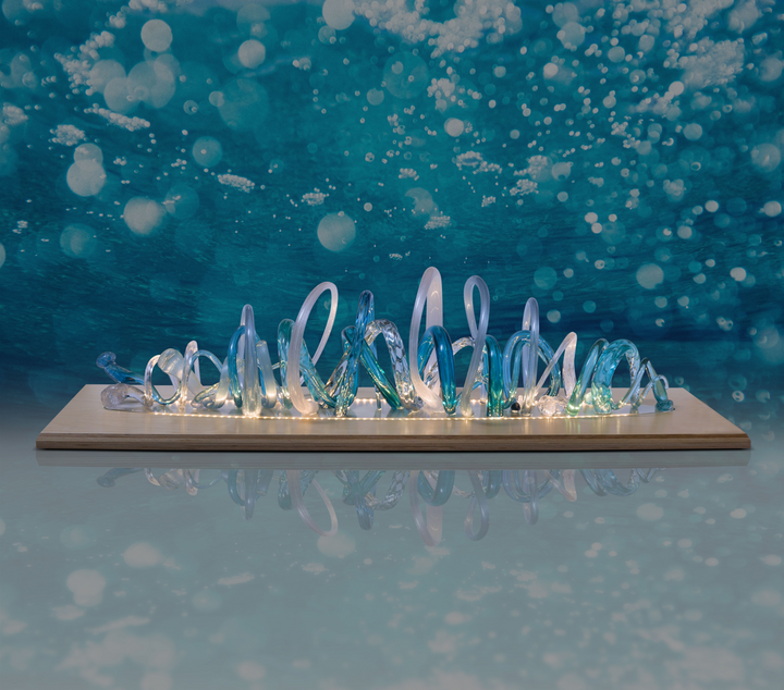 Glass sculpture of a wave, featuring flowing, ribbon-like forms in blue, aqua, and clear tones, set on a light wooden base.